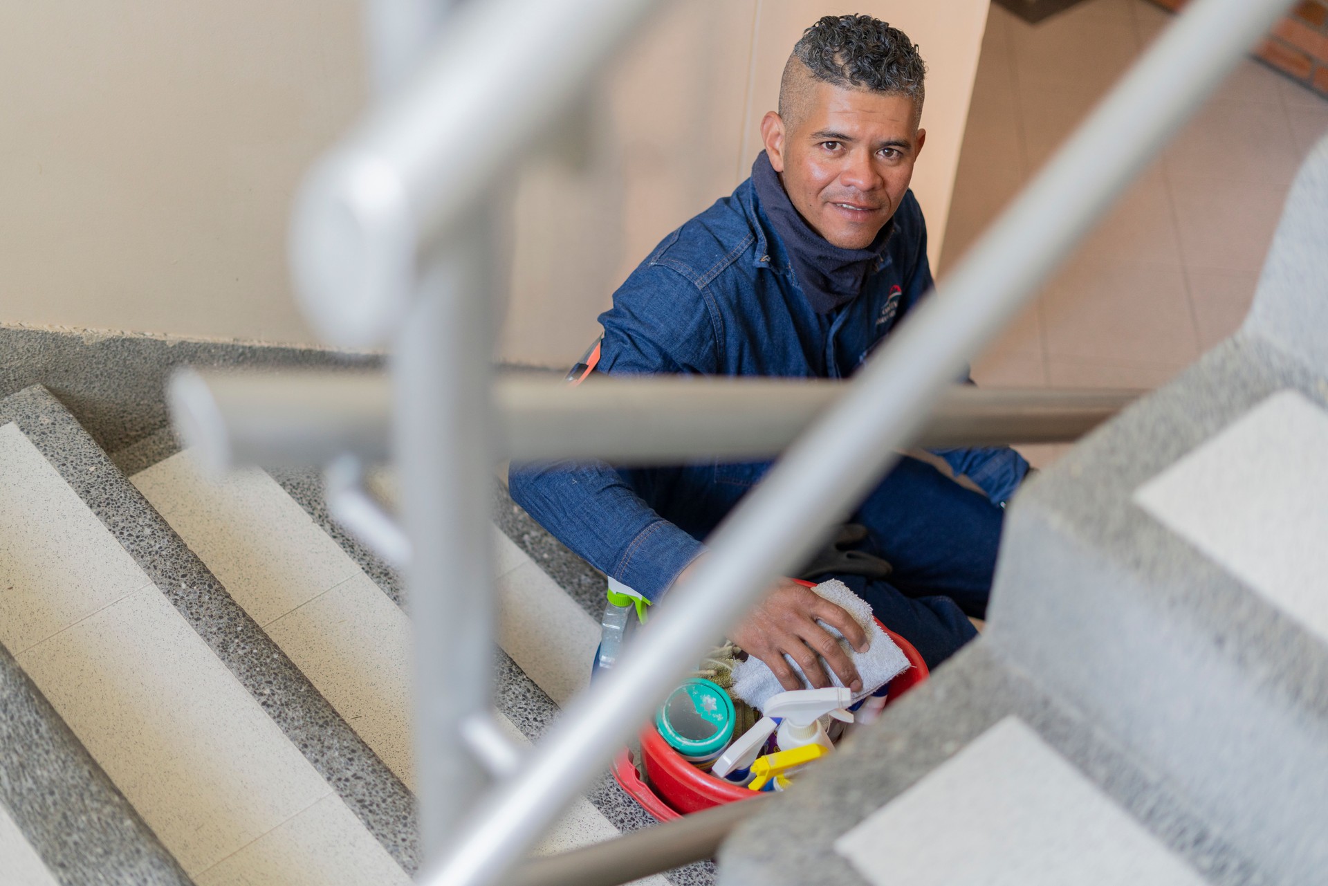 Latino man who works as a building cleaner is sitting on the stairs of the property with his cleaning products looking at the camera