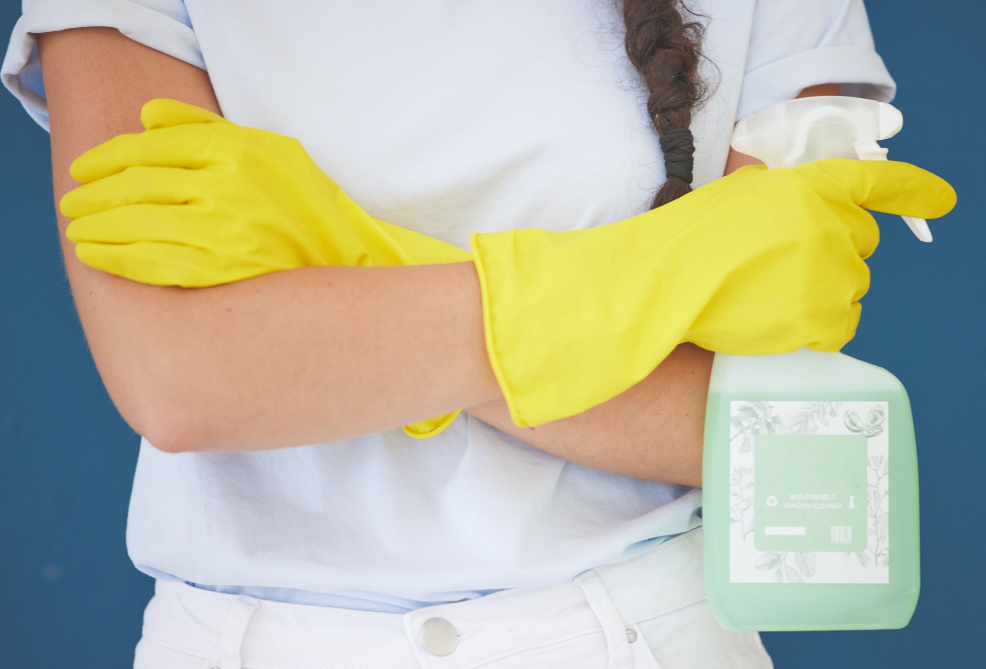 Cleaning woman and arms crossed with spray product for home hygiene and sanitation routine in blue studio. Housekeeping girl with detergent chemical bottle and safety gloves for spring cleaning.