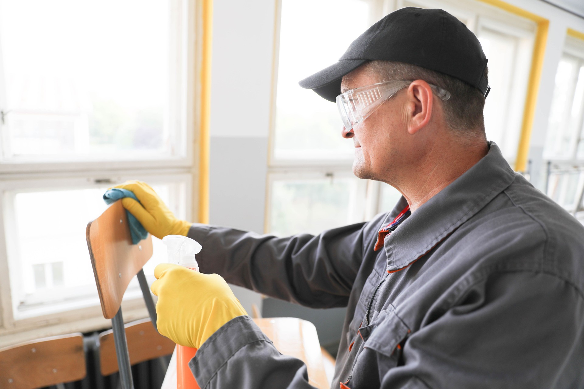 Senior Man 50+ in Work Overalls Using Handheld Pump Sprayer and Cloth to Clean Chairs in School